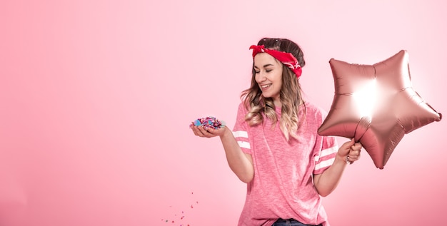 Free photo funny girl in a pink t-shirt with balloons and confetti gives a smile and emotions on a pink background