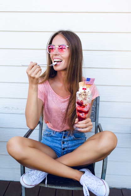 Funny girl sitting on a chair eating ice-cream with cherry on top with a spoon. She wears denim shorts, pink top and white sneakers. She has pink eyeglasses