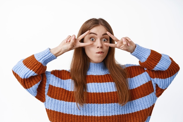 Free photo funny girl student showing peace vsigns over eyes making silly cute face having fun standing in sweater against white background copy space