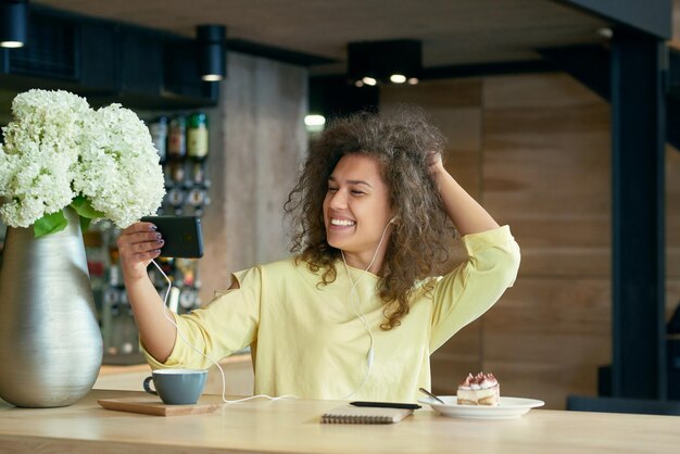 Funny girl with curly hair taking selfie sitting on restaurant's wooden table