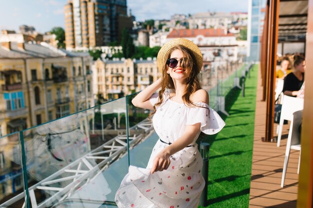 Funny girl with long hair in sunglasses is moving around on the terrace in cafe . She wears a white dress with bare shoulders, red lipstick and hat . She looks happy.