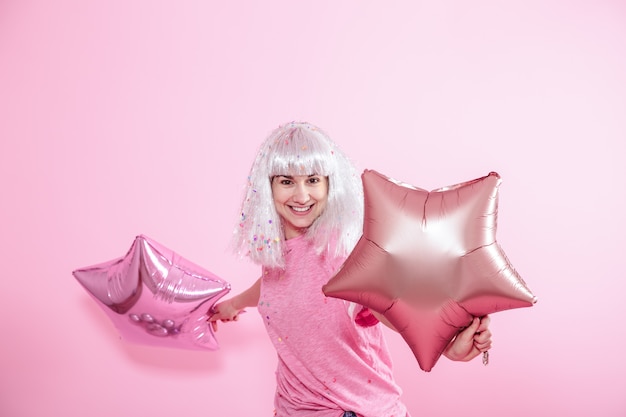 Funny Girl with silver hair gives a smile and emotion on pink background. Young woman or teen girl with balloons and confetti