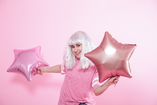 Смешная девчонка с серебряными волосами дарит улыбку и эмоции на розовом фоне. Молодая женщина или девушка с воздушными шарами и конфетти