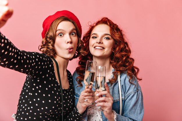 Funny girls with champagne taking selfie. Two best friends enjoying event and holding wineglasses on pink background.