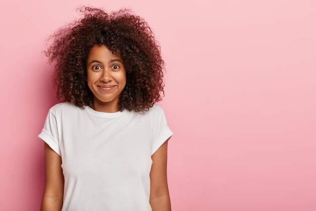 Funny good looking curly haired female teenager has pleased expression, satisfied with how things going, rejoices nice surprise or good news, wears white t shirt, models over rosy  wall