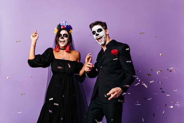 Веселый парень и темноволосая дама с раскрашенными лицами и короной из цветов позируют, танцуют в черном наряде для вечеринки.