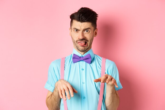 Funny guy looking at something disgusting with aversion and cringe, show tongue and shaking hands in disapproval, standing on pink background.