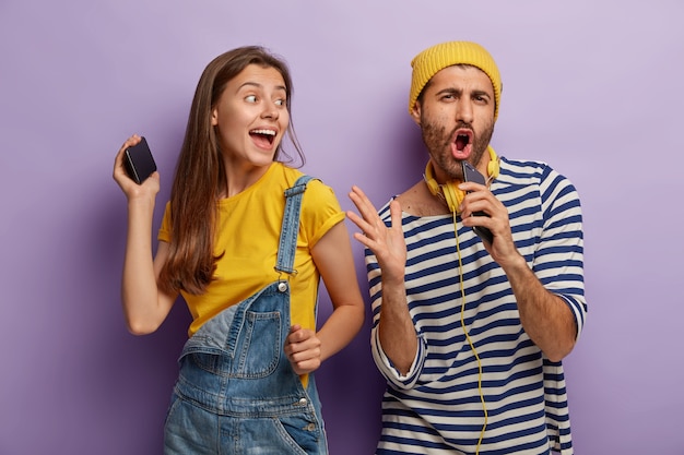 Funny guy sings favourite song, holds mobile phone near mouth as if microphone, upbeat woman dances near, have fun at party, wear fashionable clothes, have happy expressions