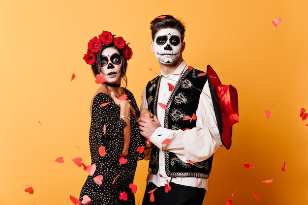 Funny guy with skeleton mask on his face in Mexican traditional vest holds his beloved hands, posing under confetti of paper hearts