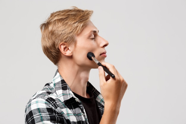 Funny handsome blond young man wearing casual plaid shirt with make-up brush over grey wall