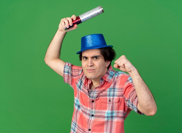 Free photo funny handsome caucasian man wearing blue party hat holds confetti cannon and keeps fist