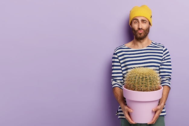 Free photo funny handsome man keeps lips folded, raises eyebrows, holds big cactus, likes growing indoor plants, dressed in casual wear