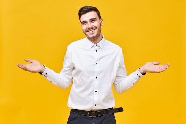 Funny handsome young Caucasian man in white shirt making helpless gesture, shrugging shoulders, being at loss, smiling, having forgetful confused facial expression, saying I don't know or Who knows