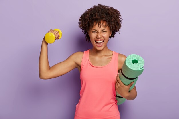 Funny happy dark skinned woman raises hand with dumbbell, shows biceps, holds rolled fitness mat