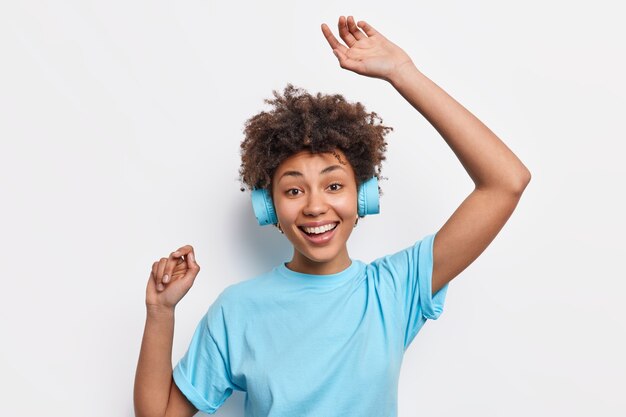 Funny happy dark skinned young Afro American woman in casual basic t shirt dances with rhythm of music wears stereo headphones isolated over white wall. People joy lifestyle hobby concept