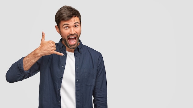 Funny happy male keeps hand near ear, makes call gesture, pretends speaking with someone, keeps mouth widely opened, dressed in fashionable shirt, isolated over white wall, free space aside.