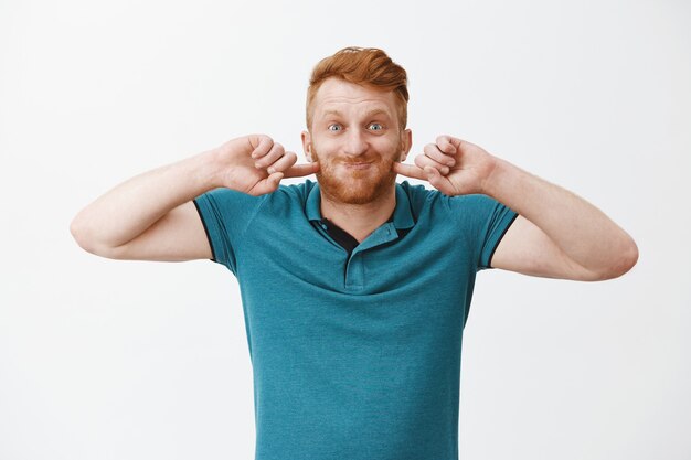 Free photo funny and happy redhead man, puffing up, holding breath and poking cheeks while smiling broadly, fooling around and making faces over gray wall