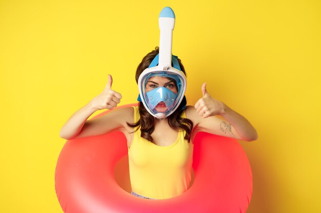 Funny happy woman in swimming ring, wearing snorkling mask for diving, showing thumbs up, good approval gesture, posing against yellow background