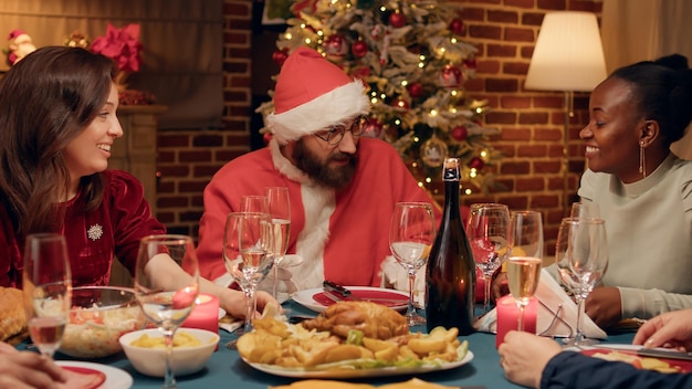 Funny husband disguised as Santa Claus discussing with people while celebrating winter holiday. Festive man wearing Santa Claus costume at Christmas dinner party talking with family members.