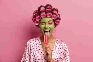 Free photo funny joyful housewife licks delicious ice cream, winks eye, gets pleasure from eating cold refreshing summer dessert, wears green mask on face, hair rollers, dressed in casual outfit, has fun at home