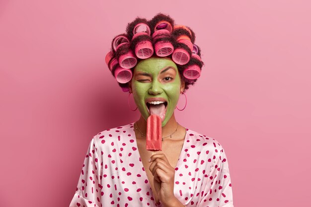 Funny joyful housewife licks delicious ice cream, winks eye, gets pleasure from eating cold refreshing summer dessert, wears green mask on face, hair rollers, dressed in casual outfit, has fun at home