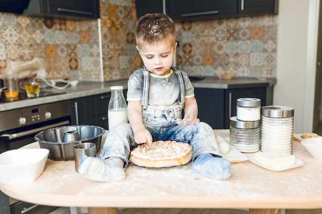 Free photo funny kid sitting on the kitchen table in a roustic kitchen playing with flour and tasting a cake.