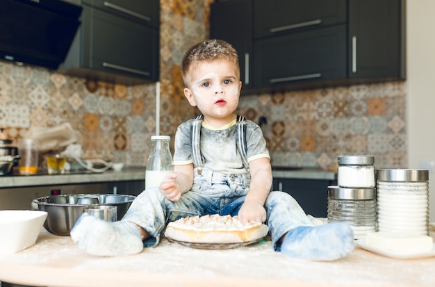 Funny kid sitting on the kitchen table in a roustic kitchen playing with flour and tasting a cake.