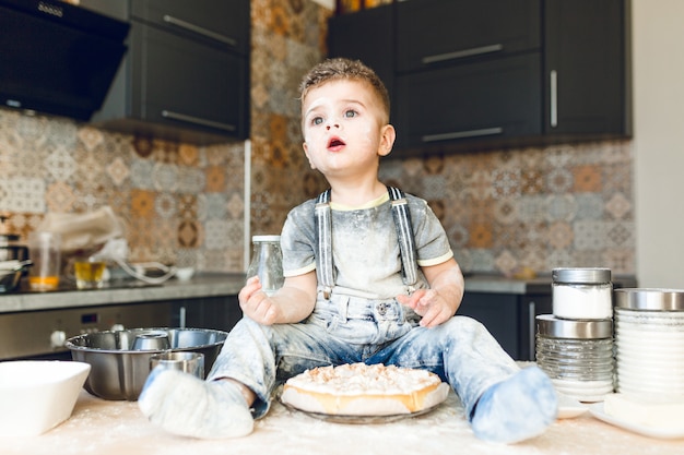 Bambino divertente che si siede sul tavolo della cucina in una cucina rustica che gioca con la farina e assaggia una torta.