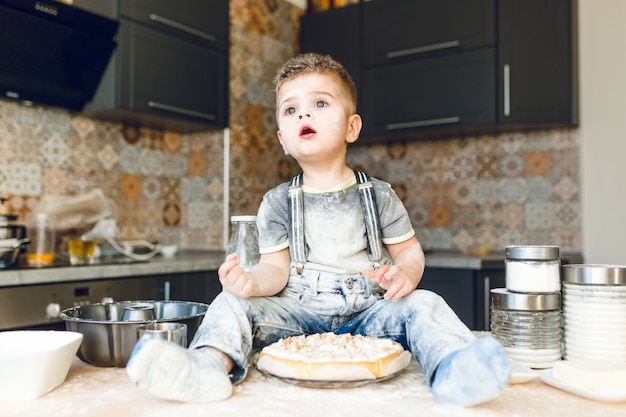 Funny kid sitting on the kitchen table in a roustic kitchen playing with flour and tasting a cake.