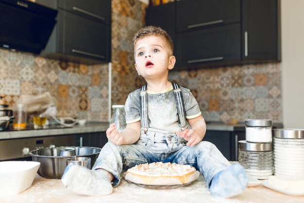 Funny kid sitting on the kitchen table in a roustic kitchen playing with flour and tasting a cake.