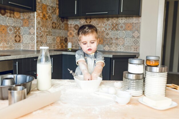 Funny kid standing in a roustic kitchen playing with flour. He is covered in flour and looks funny. He is cute. Milk and different jars stand on the table.