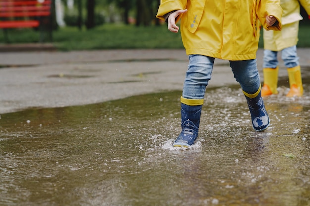 Funny kids in rain boots playing with paper ship by a puddle