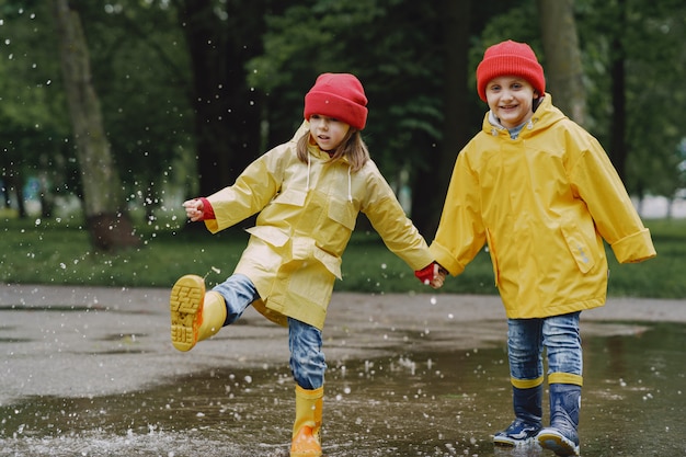 Funny kids in rain boots playing with paper ship by a puddle