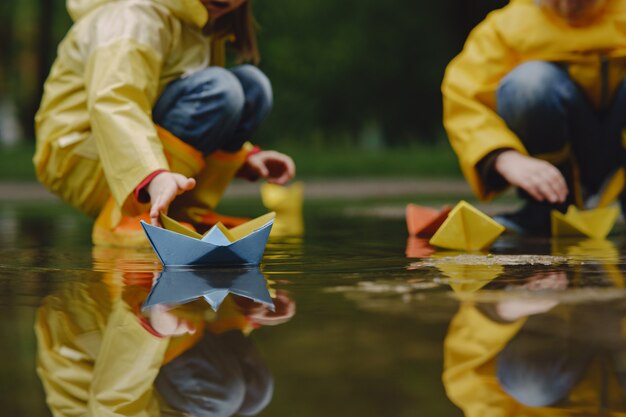 Funny kids in rain boots playing with paper ship by a puddle