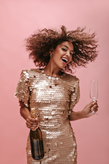 Funny lady in stylish beige dress holding bottle with champagne and glass and playing her curly fluffy hair on isolated wall..