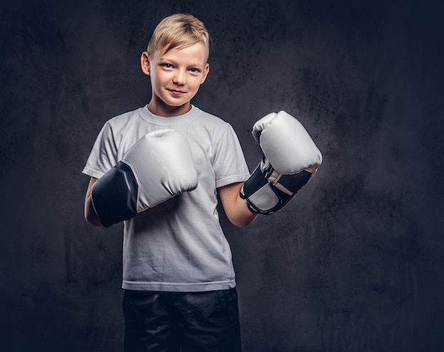 Funny little boy boxer with blonde hair dressed in a white t-shirt wearing boxing gloves posing in a studio. Isolated on a dark textured background.