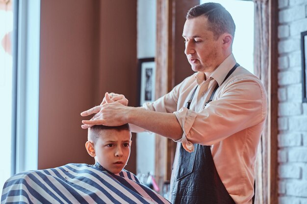 Забавный маленький мальчик впервые делает модную стрижку у опытного парикмахера в модной парикмахерской.