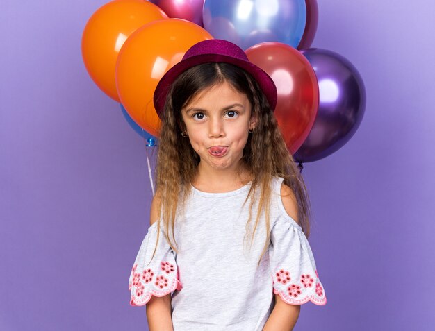 смешная маленькая кавказская девочка в фиолетовой шляпе торчит из языка, стоя перед гелиевыми шарами, изолированными на фиолетовой стене с копией пространства