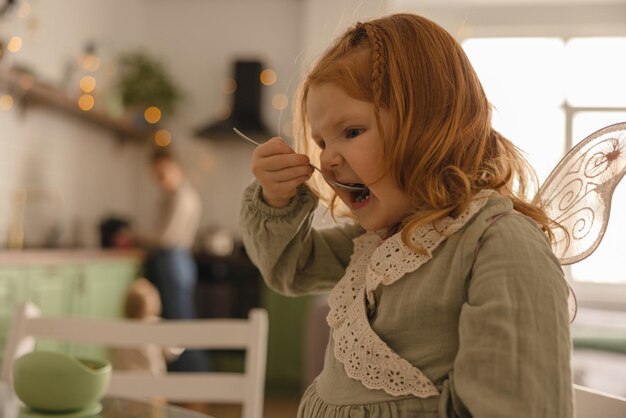 面白い小さな白人の赤毛の女の子の子供は、キッチンでスプーンで食べて幸せです子供と栄養の概念