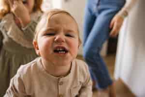 Free photo funny little fairskinned blond boy 12 years old in jacket grimaces at camera against background of his sister and mother in room fun concept with family