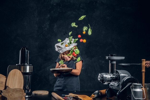 Funny little girl is tossing vegetables on the pan at dark photo studio.