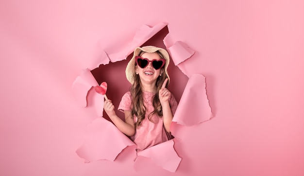 Funny little girl looks out from the hole in a beach hat and glasses in the shape of a heart, holding a heart on a stick, on a colored background, a place for text, studio shooting