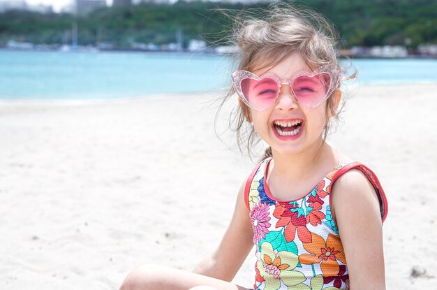 Funny little girl posing in sunglasses playing with sand on the beach. Summer entertainment and recreation.