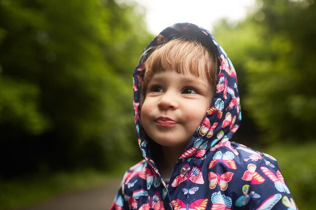 Funny little girl in rain coat stands in green park