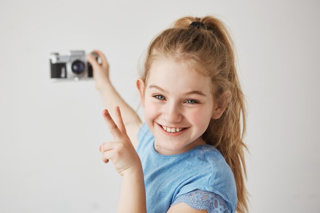 Funny little girl with blue eyes and light hair smiles, holding photocamera in her hand, showing v-sign, going to take selfie.