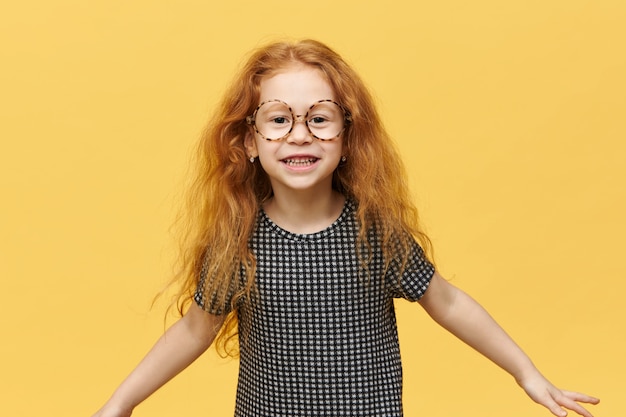 Funny little girl with long loose red hair jumping expressing true positive emotions smiling broadly wearing large round spectacles. Picture of cute cheerful child having fun posing isolated