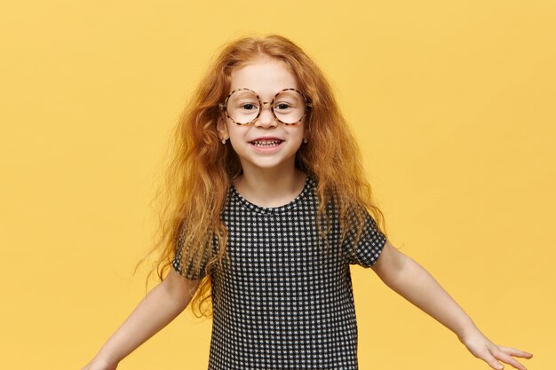 Funny little girl with long loose red hair jumping expressing true positive emotions smiling broadly wearing large round spectacles. Picture of cute cheerful child having fun posing isolated