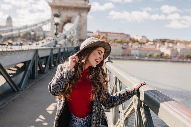 晴れた日の橋での写真撮影中に目を閉じてポーズをとって帽子をかぶった面白い長髪の女性