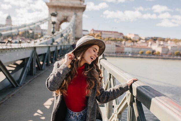 Funny long-haired woman in hat posing with eyes closed during photoshoot on bridge in sunny day