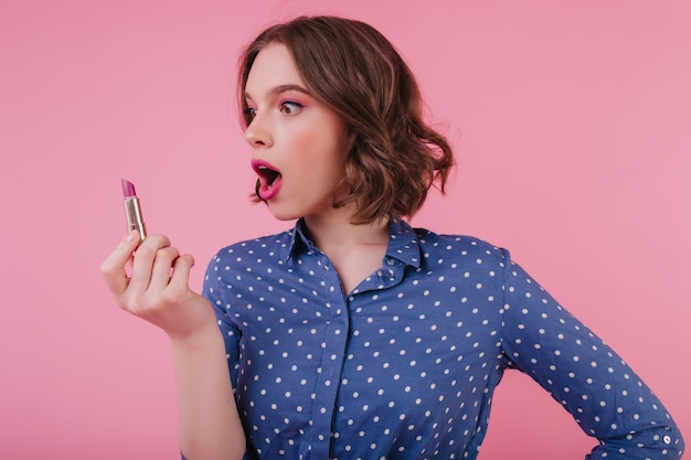 Funny lovely girl with wavy hairstyle looking at lipstick. Indoor photo of elegant female model in blue blouse doing makeup on pink wall.